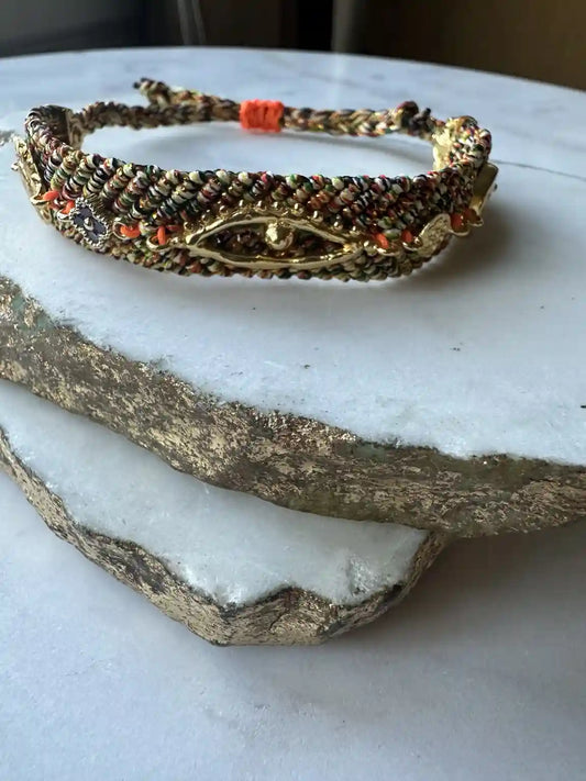 Handmade orange and blue woven cord bracelet with gold-plated evil eye charm and zircon on a textured surface