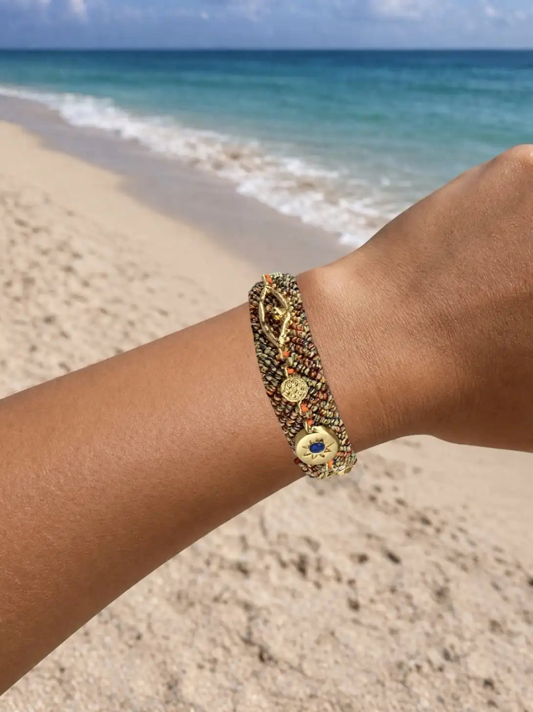 Orange, Gold, and blue woven cord bracelet with a gold charm and zircon gemstones on a person's wrist against a beach background