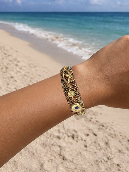 Orange, Gold, and blue woven cord bracelet with a gold charm and zircon gemstones on a person's wrist against a beach background