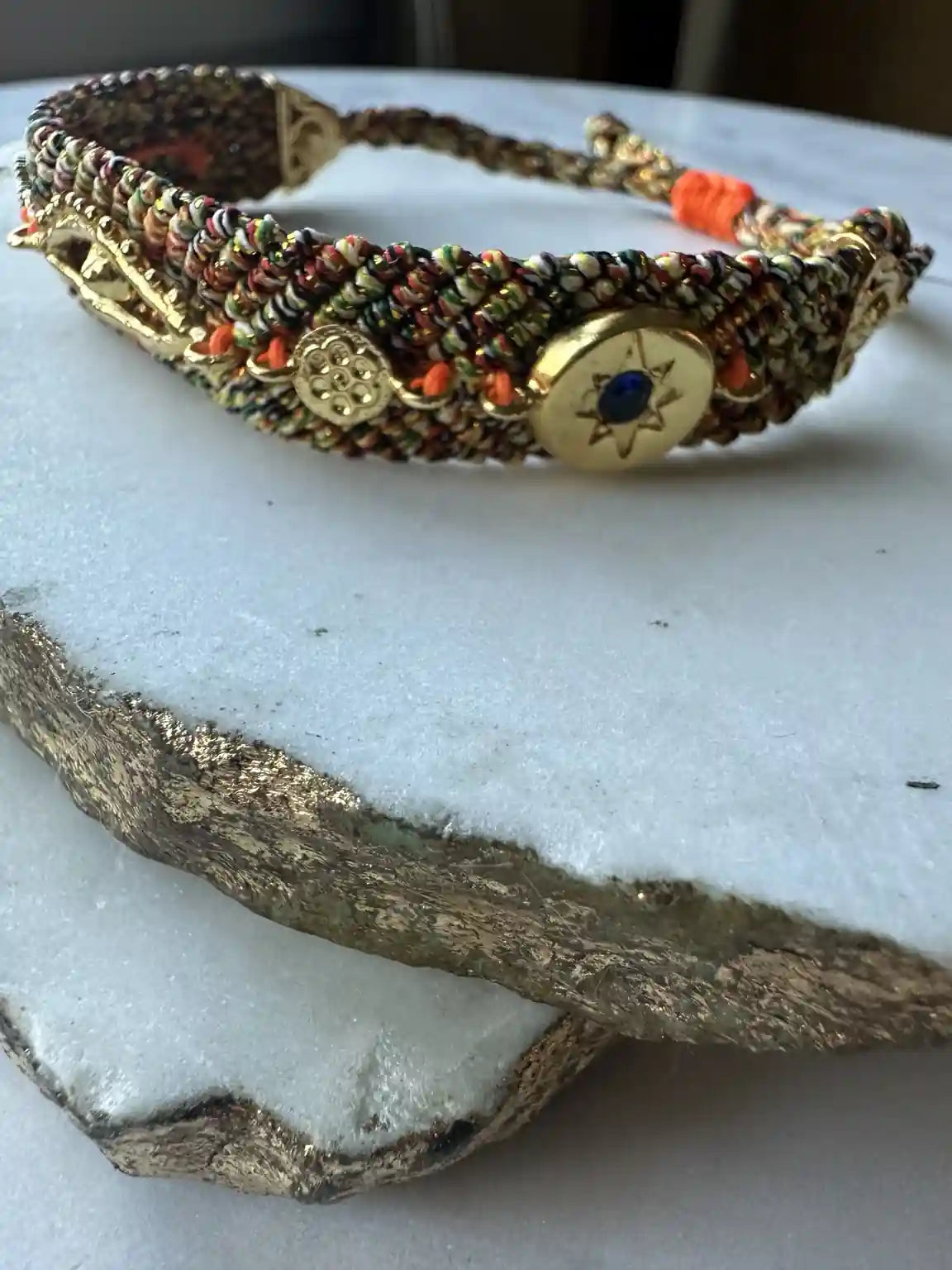 Handmade woven orange and blue cord bracelet with gold evil eye charm and zircons on a textured surface