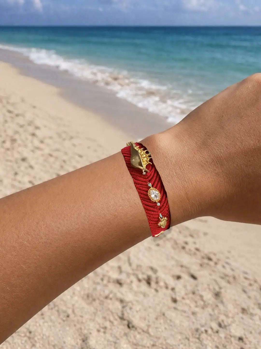 Red handmade woven bracelet with gold evil eye and beach zircon accents on a wrist against a beach background