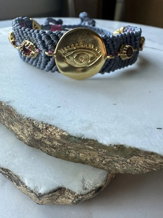 Slate gray woven cord bracelet with gold-played evil eye charm and zircon on a textured surface
