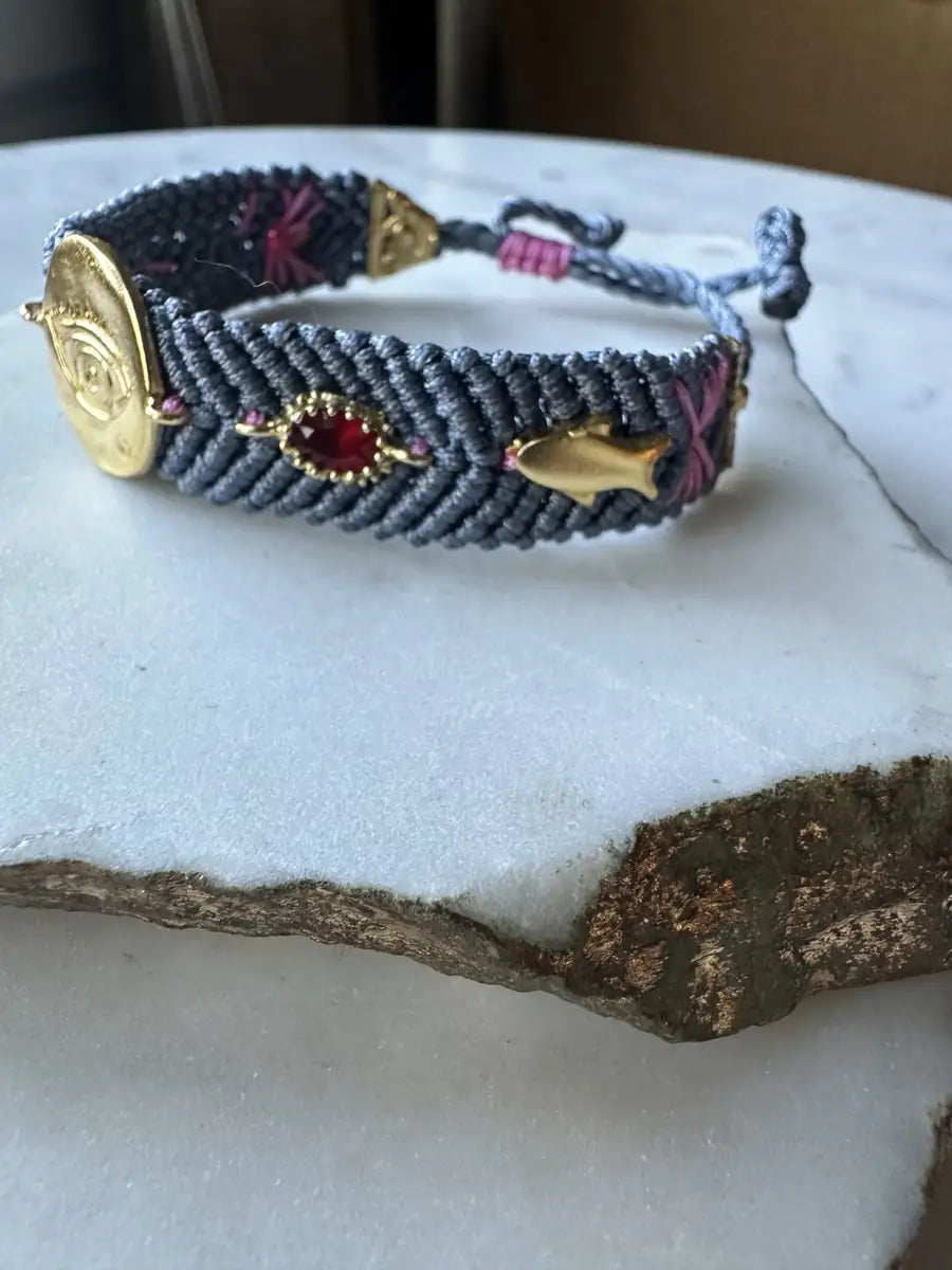 Slate gray handmade woven cord Bracelet with gold evil eye charm and cubic zircon charms on a textured surface