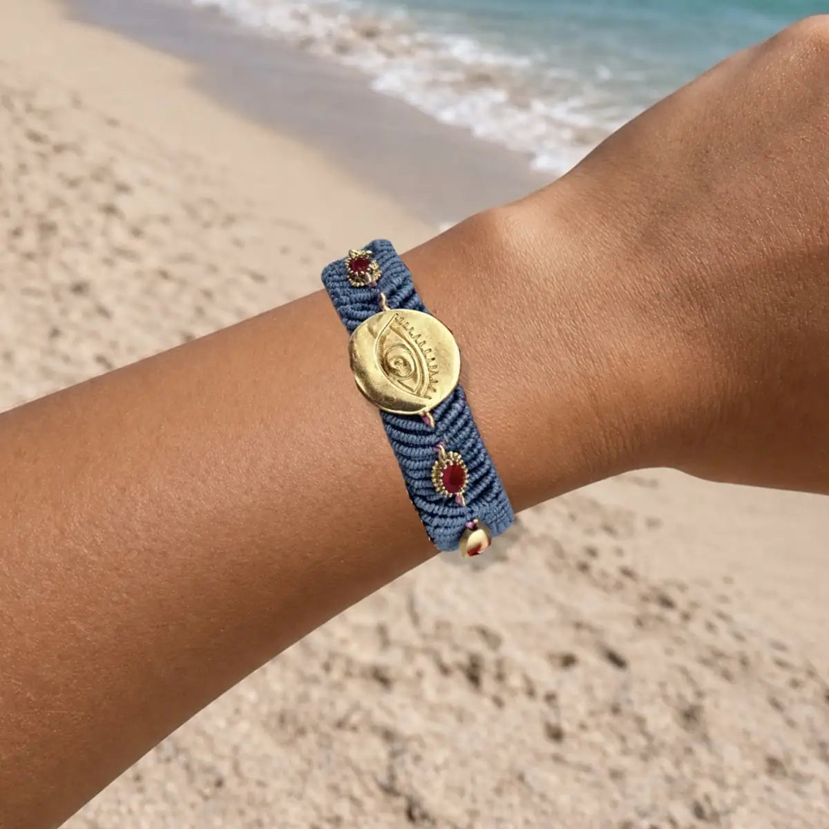 Slate gray woven cord bracelet with a gold evil eye charm and zircon on a wrist against a beach background