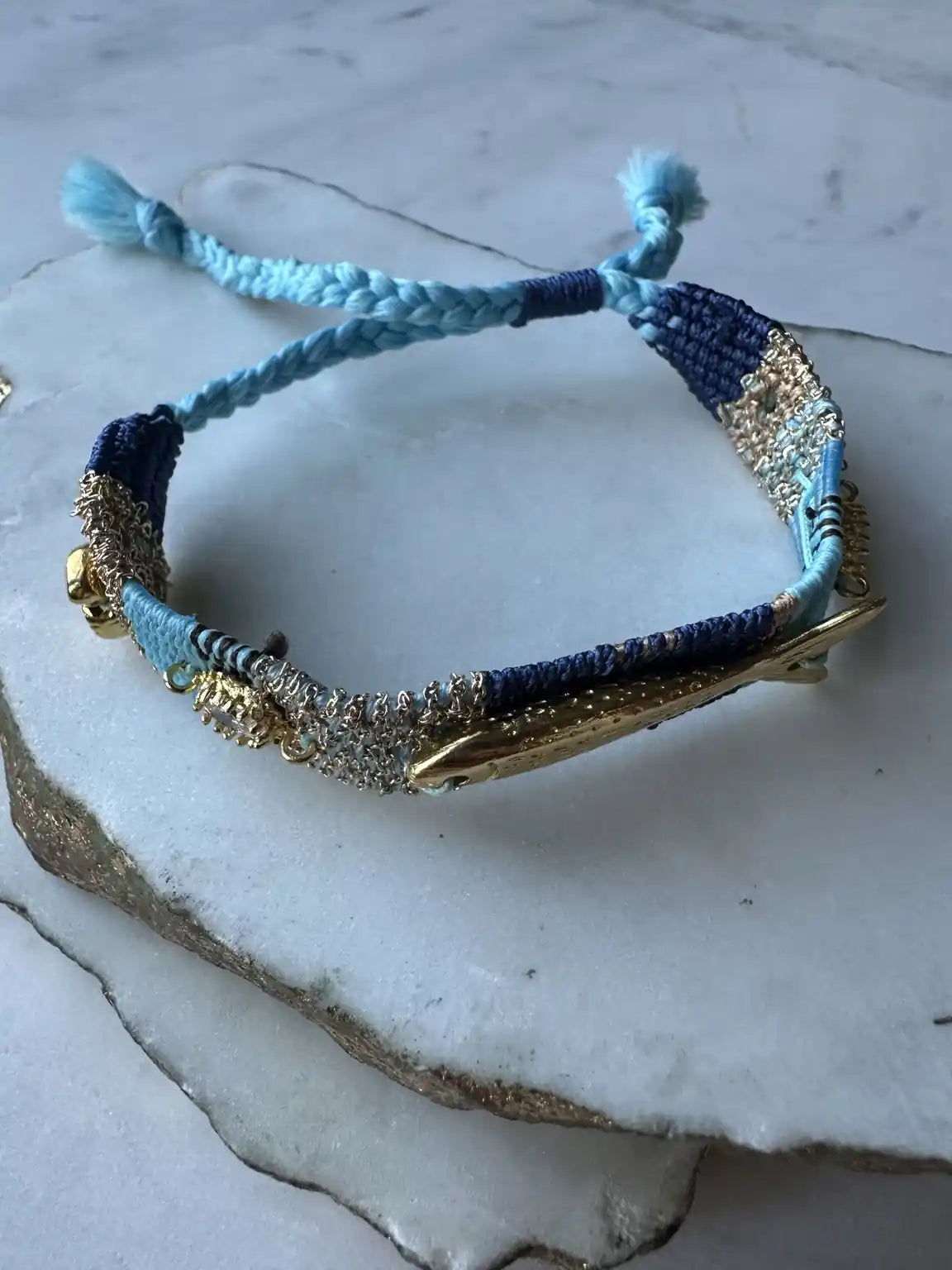 Blue, turquoise, and gold braided handmade woven bracelet with gold-plated fish charm and beach charms and crystals on a marble surface
