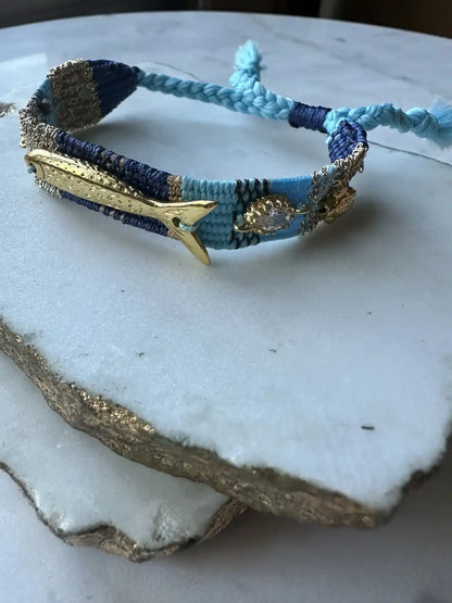 Handmade blue, turquoise and gold woven Bracelet with gold-plated fish and beachwear charms and crystals on a marble surface