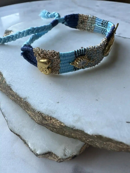 Turquoise, Blue and gold hand woven bracelet with gold-plated fish charm and beachwear charms and crystals on a textured white surface