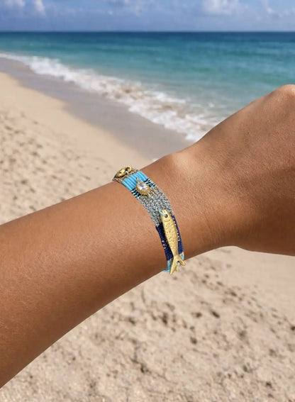 Blue, turquoise and gold handmade woven Bracelet with gold-plated fish charm on wrist against a beach background
