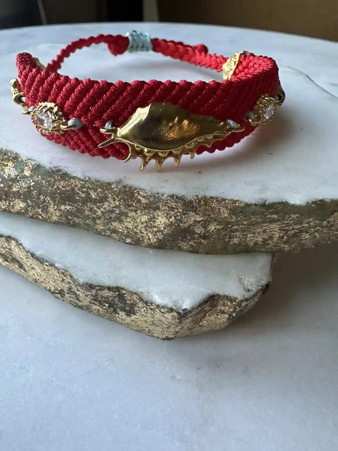 Handmade Red woven bracelet with gold-plated shell and evil eye accents on a marble surface