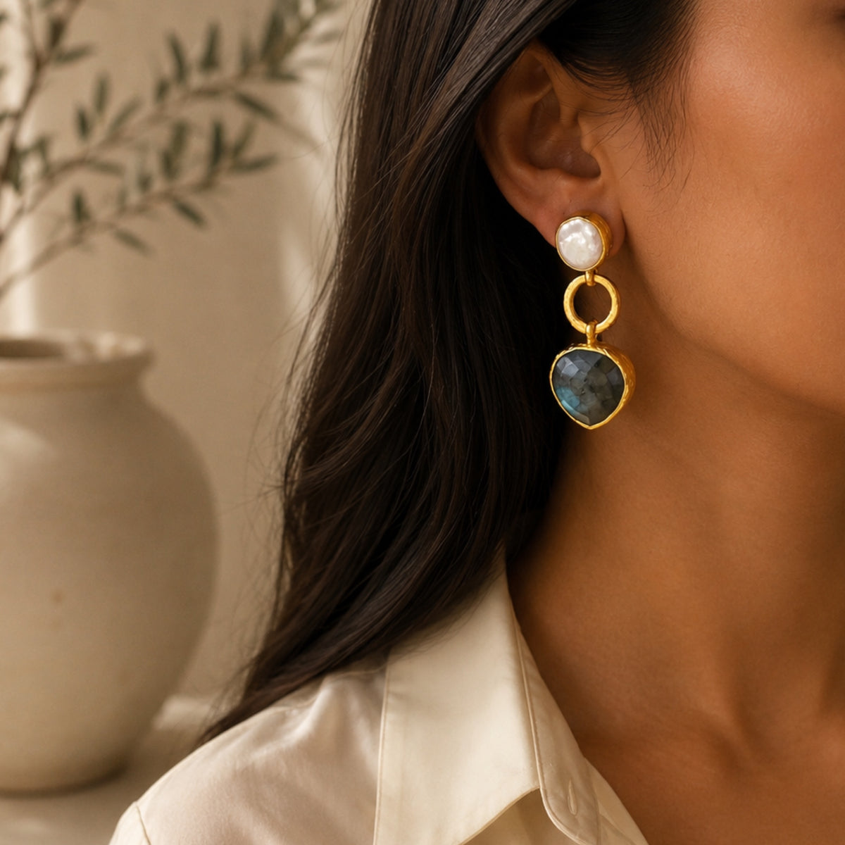 A close-up of a woman wearing a contemporary gold dangle earring with a white flat pearl stud with labradorite stone, set against a softly blurred indoor background with a ceramic vase.