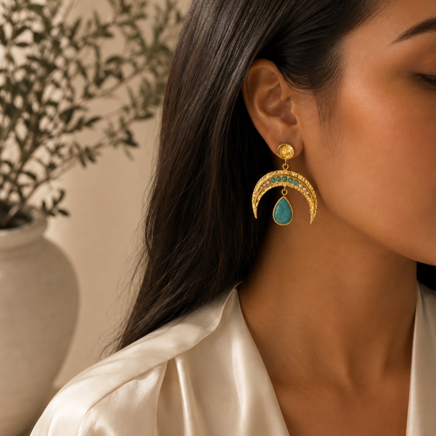 A close-up lifestyle shot of a woman wearing a gold crescent moon dangle earring with turquoise blue stones and pearls and a teardrop-shaped turquoise center, set against a soft beige background with a blurred plant.