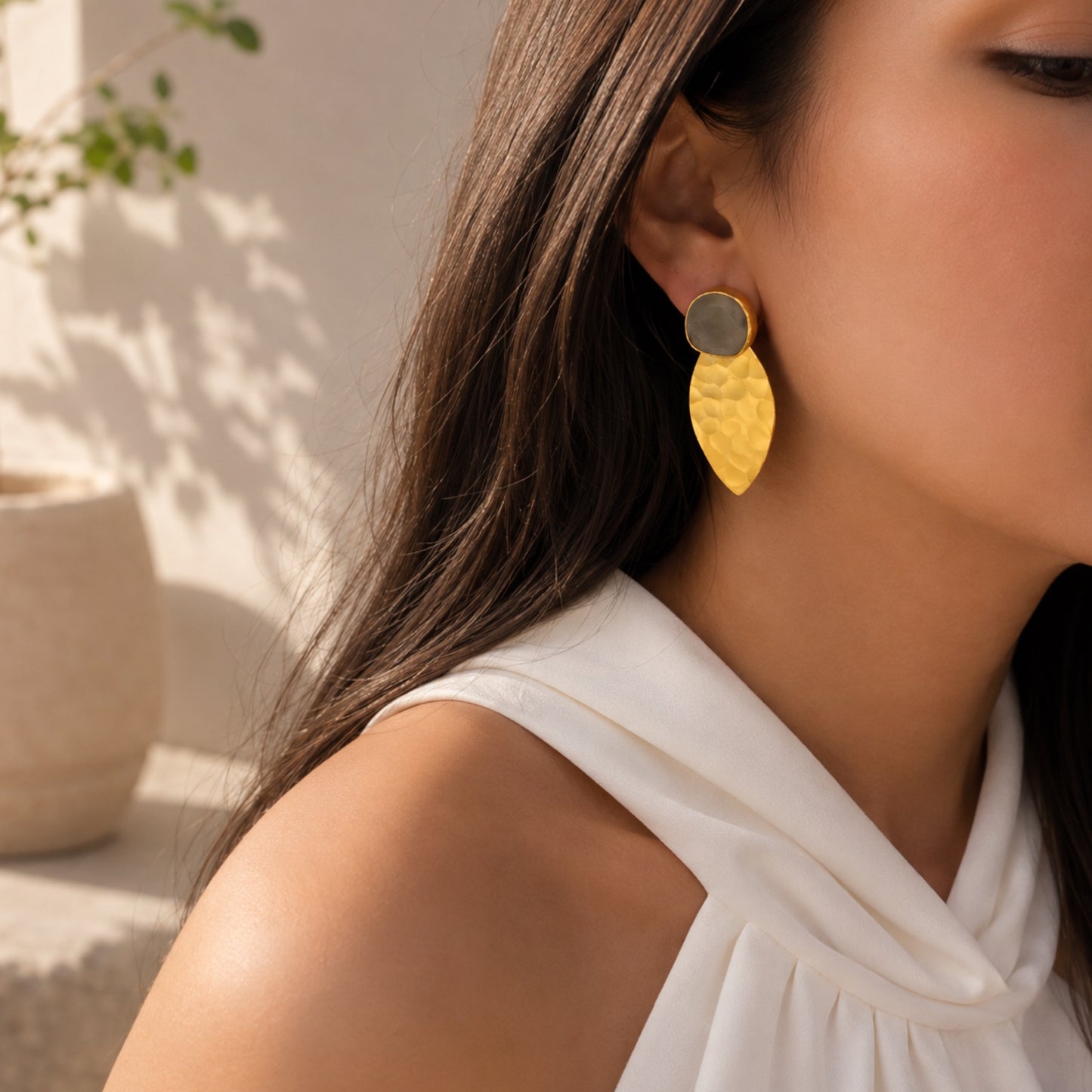 A close-up lifestyle shot of a woman wearing a gold leaf-shaped drop earring with a textured hammered finish and a round gray gemstone