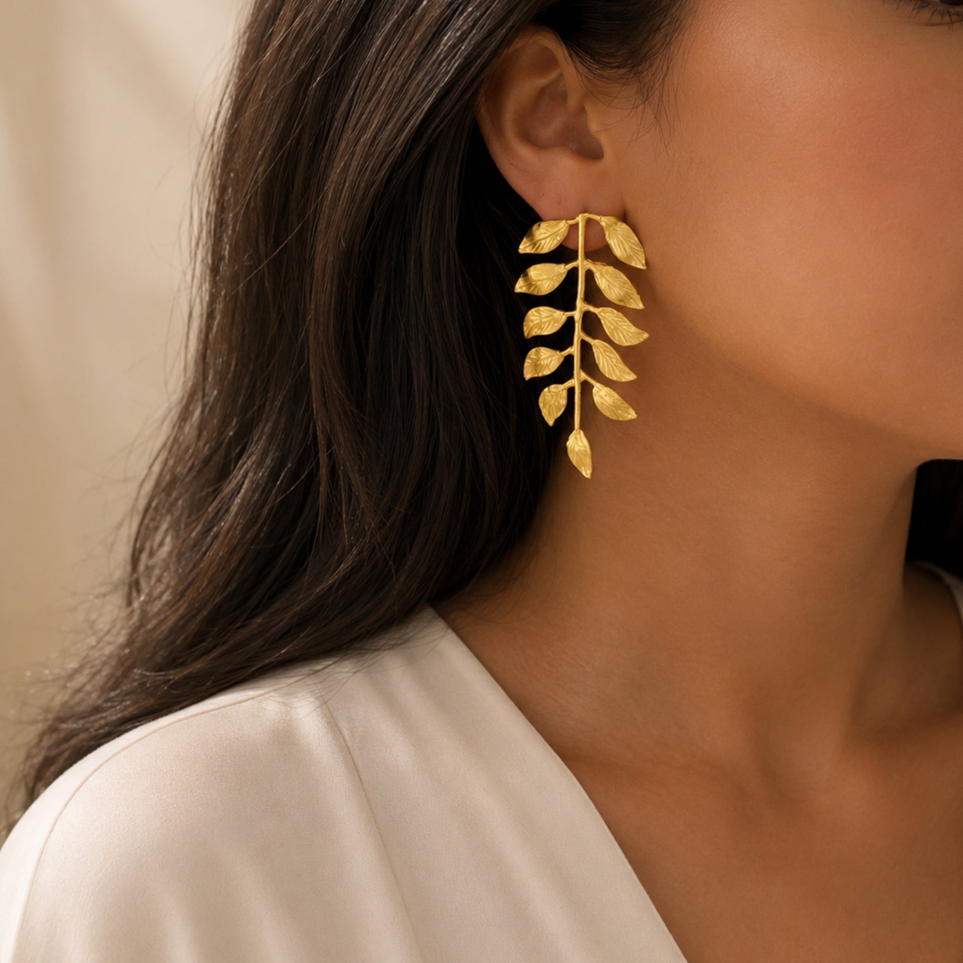 A close-up of a woman wearing a gold vine leaf drop earring, showing the textured gold leaf design against her ear and neck.