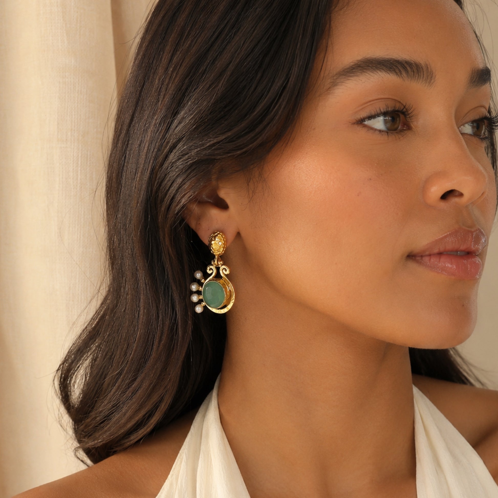 A close-up of a woman wearing a gold handmade aqua chalcedony and pearl statement earring with neutral backdrop 