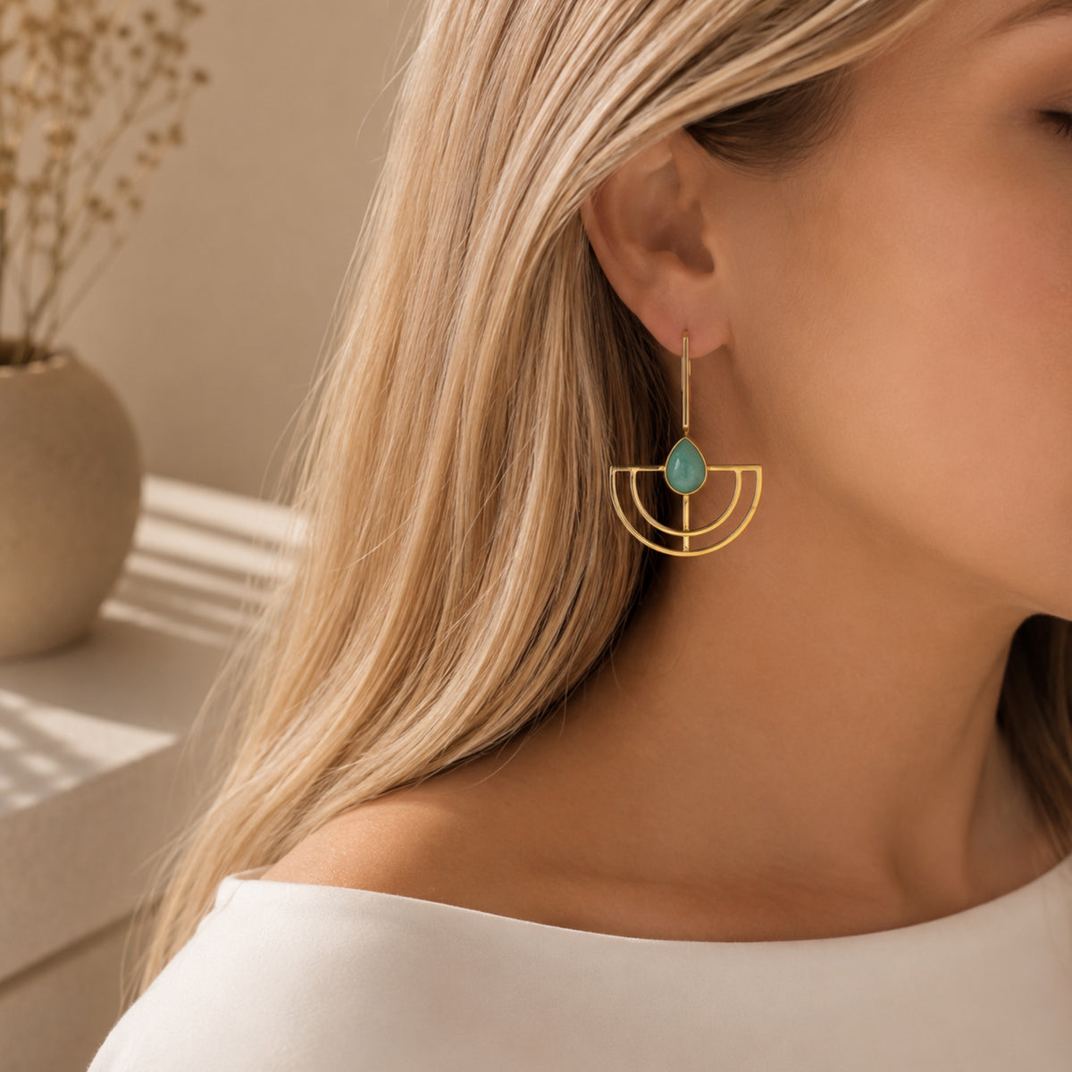 A close-up lifestyle shot of a woman wearing a gold geometric dangle earring with aqua chalcedony teardrop stone, set against a warm, softly lit background.