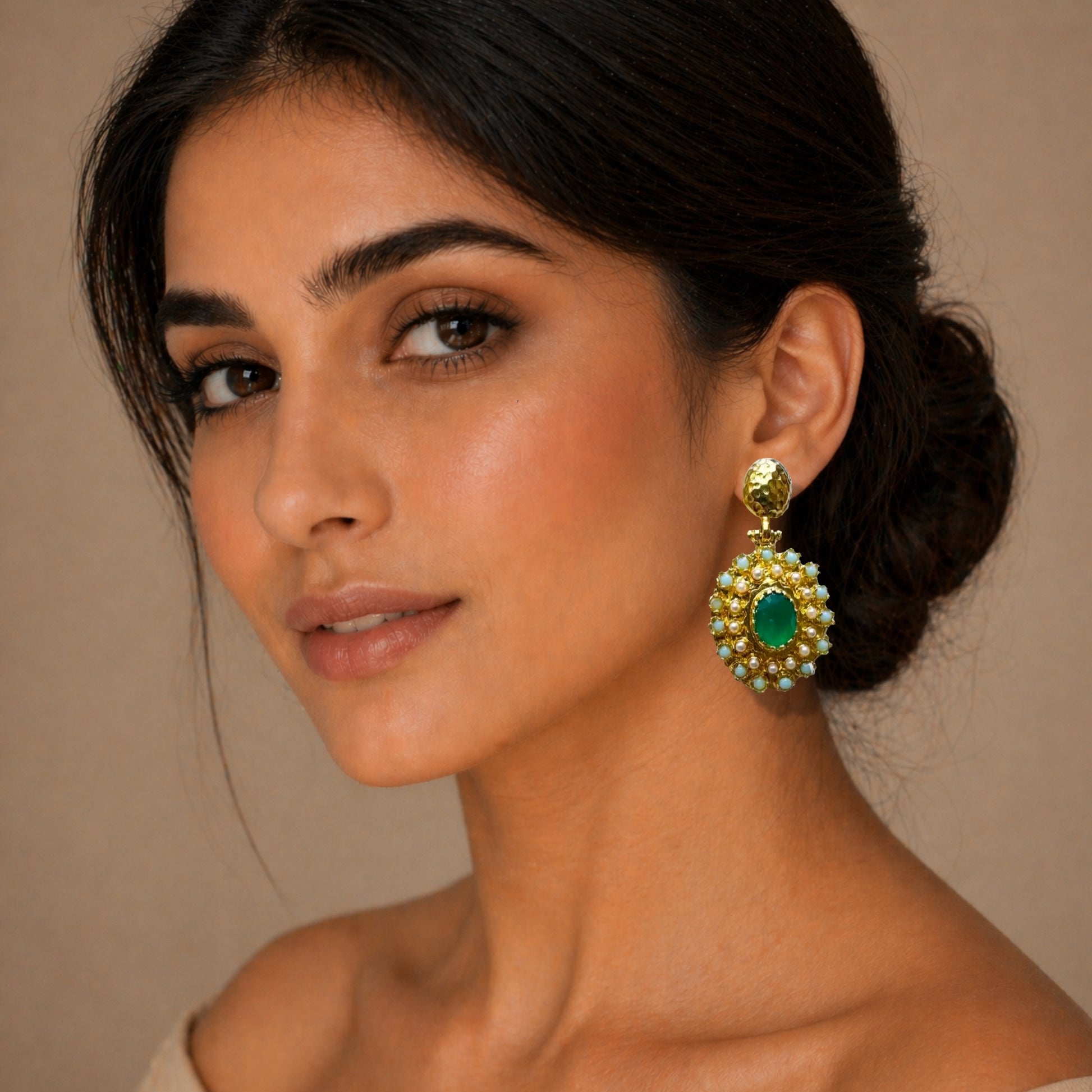A close-up of a woman wearing handmade  gold-plated statement earrings with an emerald center and surrounding white stones, against a warm beige background.