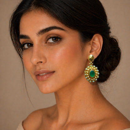 A close-up of a woman wearing handmade  gold-plated statement earrings with an emerald center and surrounding white stones, against a warm beige background.