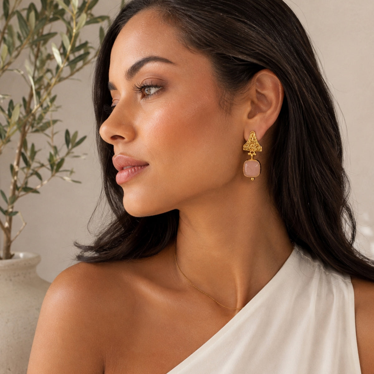 A woman in profile wearing a gold-plated statement earring with a rose quartz gemstone, against a soft beige background with a blurred olive plant.