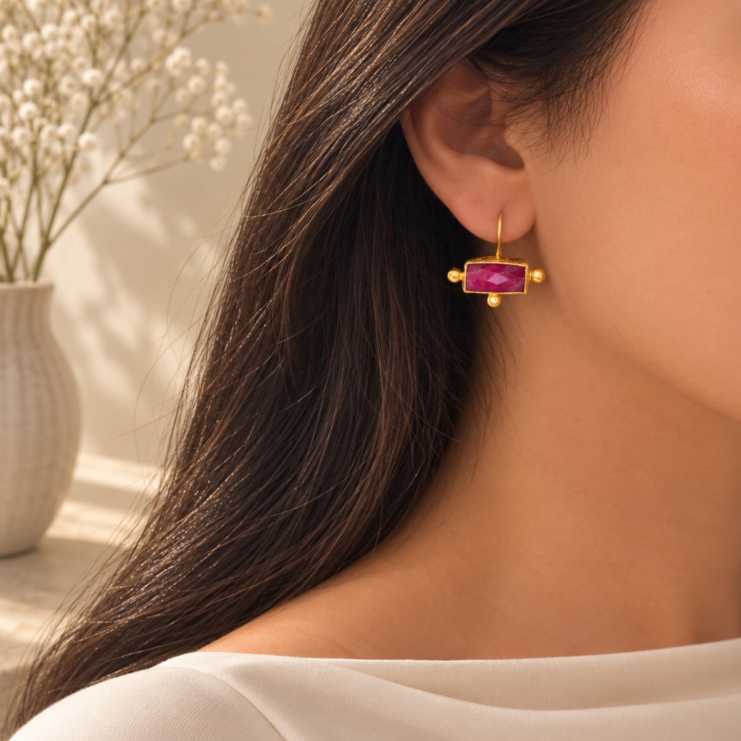 A close-up lifestyle image of a woman wearing a gold rectangle ruby drop earring against a warm, softly lit background with a blurred vase of baby's breath