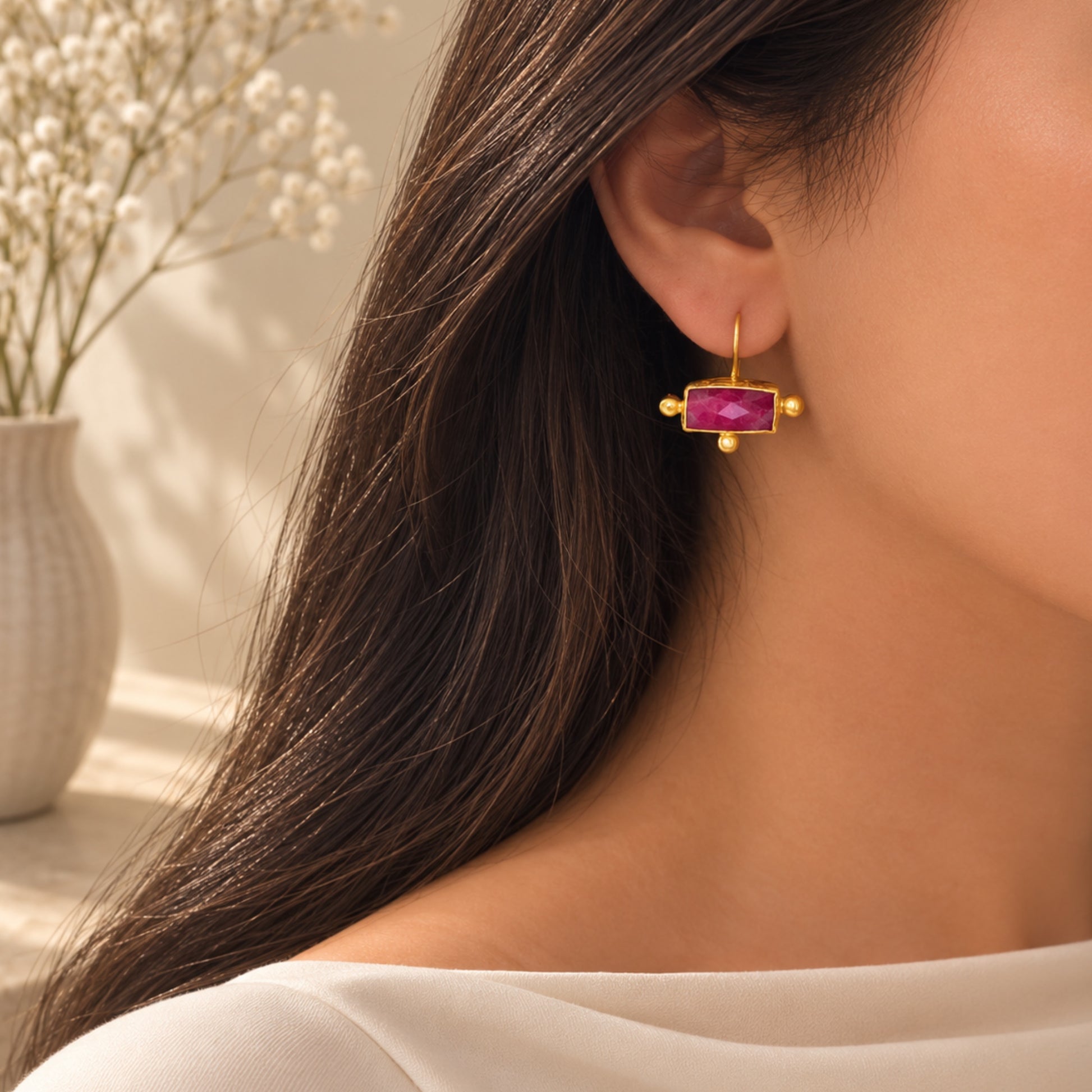 A close-up lifestyle image of a woman wearing a gold rectangle ruby drop earring against a warm, softly lit background with a blurred vase of baby's breath