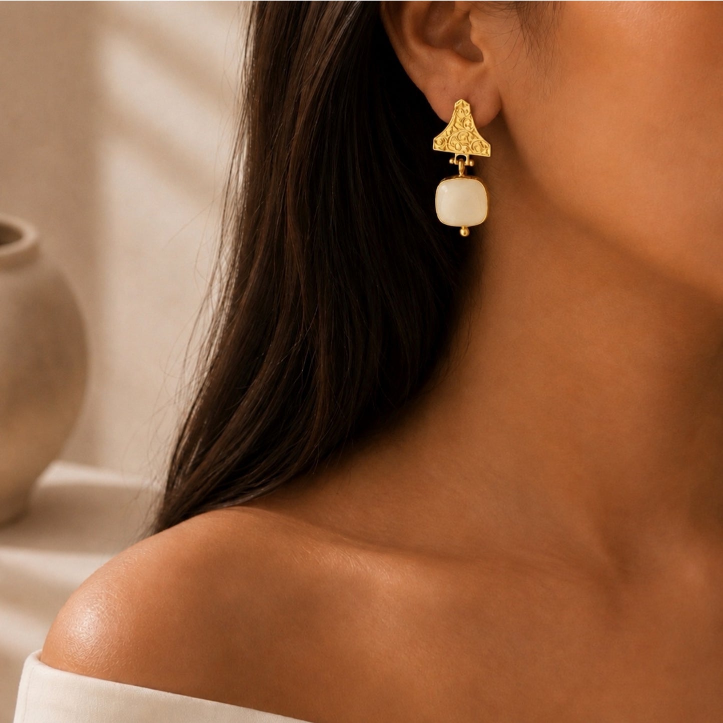 close-up of a woman wearing a gold dangle white snow Quartz gemstone earring with a, shown against a warm neutral background