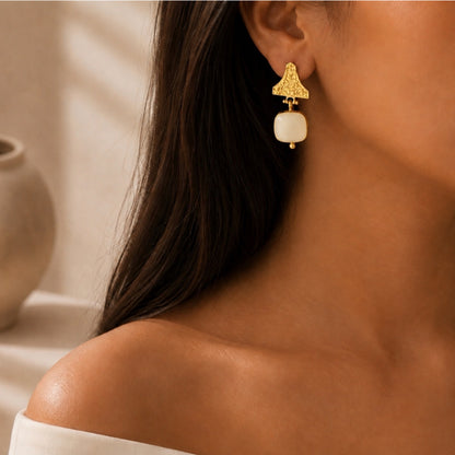 close-up of a woman wearing a gold dangle white snow Quartz gemstone earring with a, shown against a warm neutral background