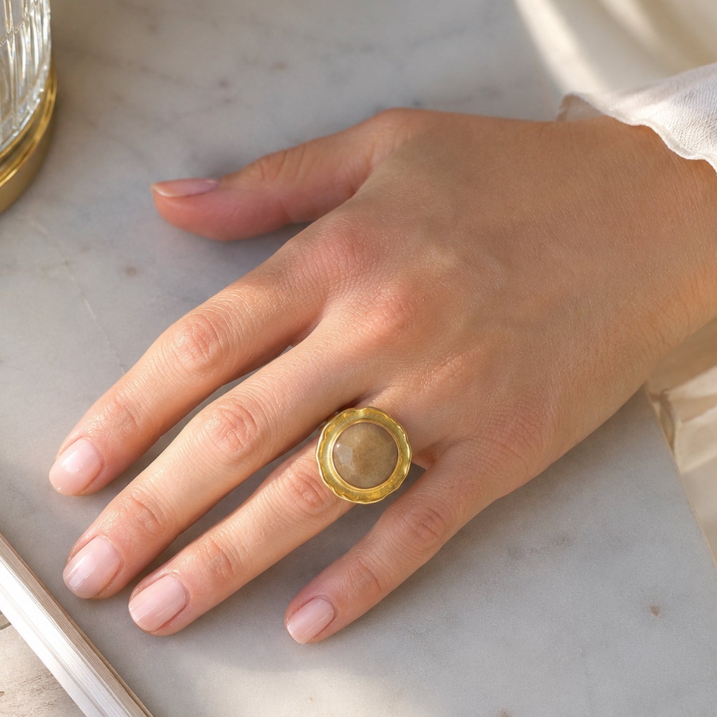 A close-up of a hand wearing a handmade gold-plated cocktail ring with a round, faceted beige quartz stone, resting on a light marble surface.