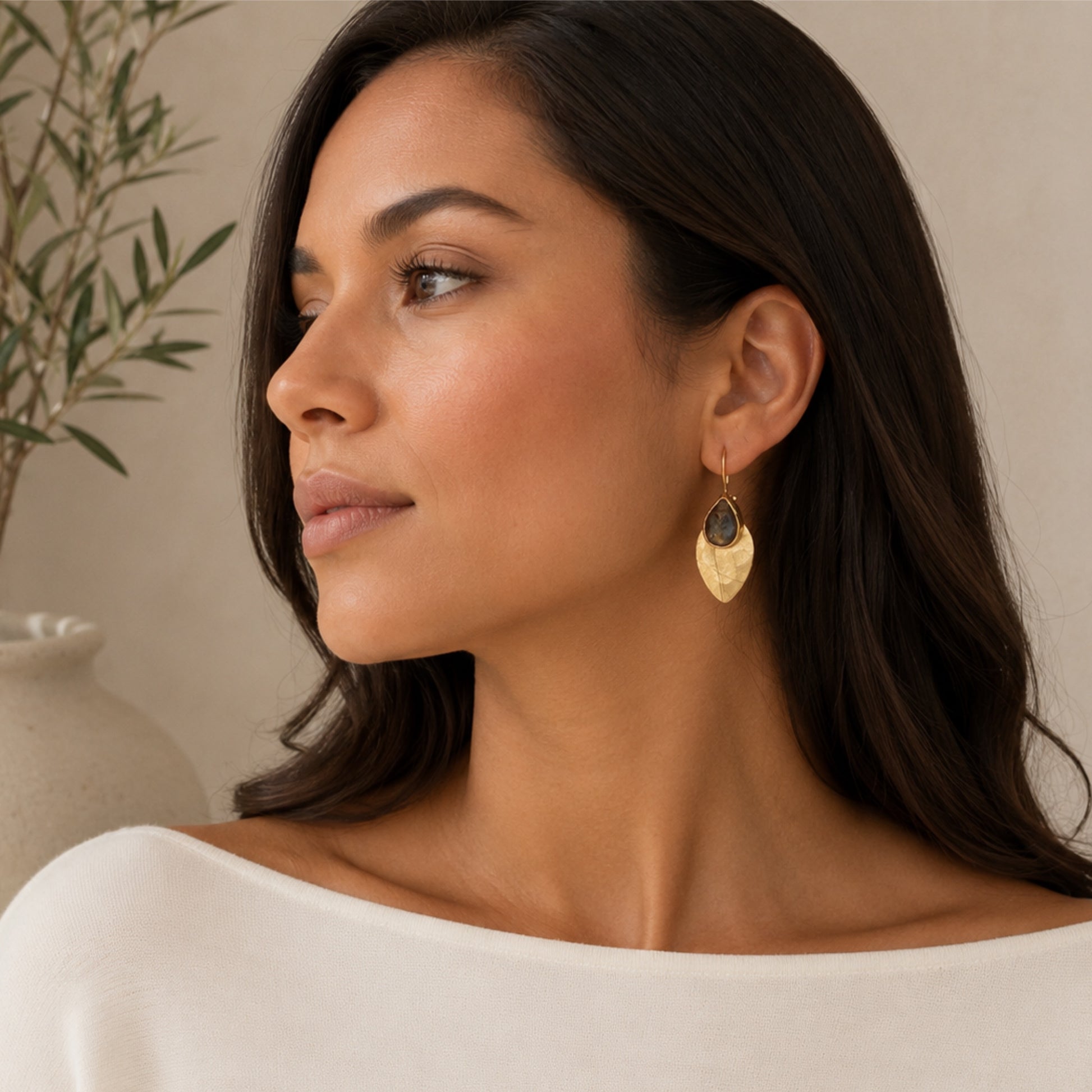 A woman in a white top wearing a gold leaf-shaped dangle earring with a labradorite gemstone, shown in a warm, softly lit indoor setting.