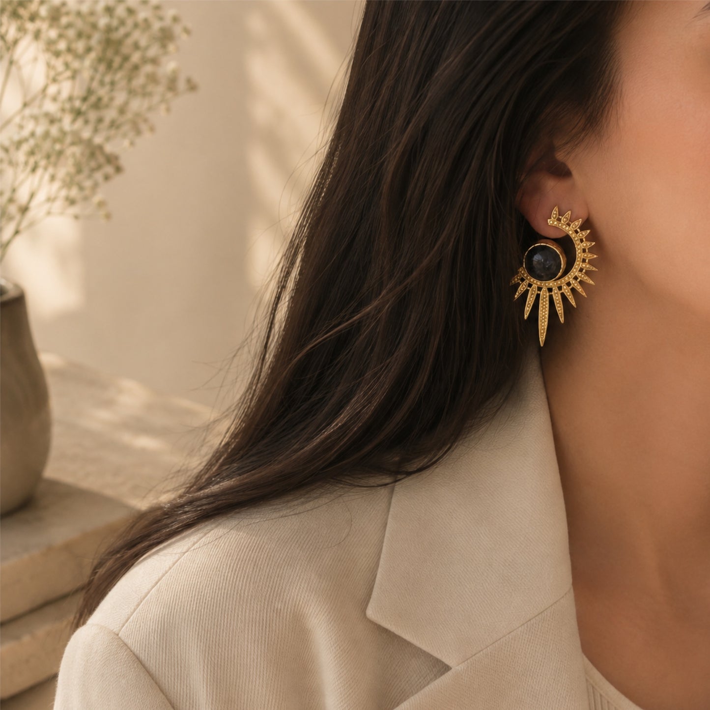 A close-up lifestyle shot of a woman wearing a gold abstract earring with a blue lapis lazuli circular center, styled with a beige blazer in warm, natural light.