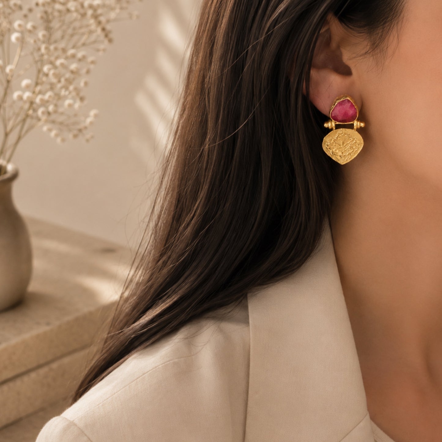 A close-up lifestyle shot of a woman wearing a gold ruby stud earring against a warm beige background with soft shadows