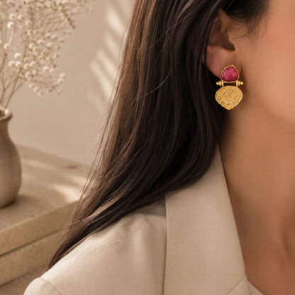 A close-up lifestyle shot of a woman wearing a gold ruby stud earring against a warm beige background with soft shadows