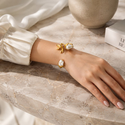 A close-up lifestyle shot of a woman's hand and wrist wearing handmade gold-plated cuff bangle with white baroque pearls and floral design resting on a beige marble surface.