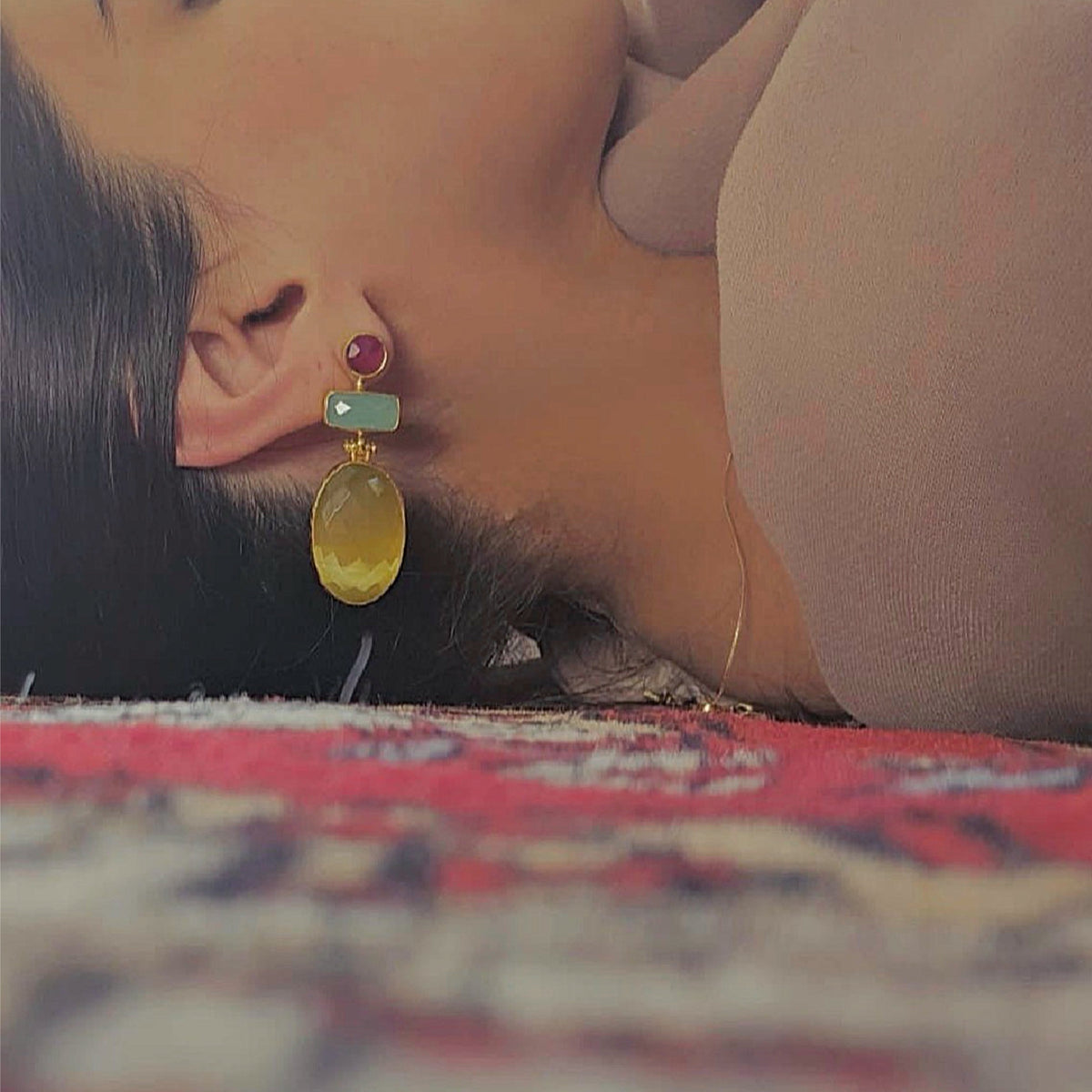 Model laying in floor wearing A pair of gold drop earrings featuring a round ruby gemstone, a rectangular aqua chalcedony stone, and a large faceted oval yellow agate stone