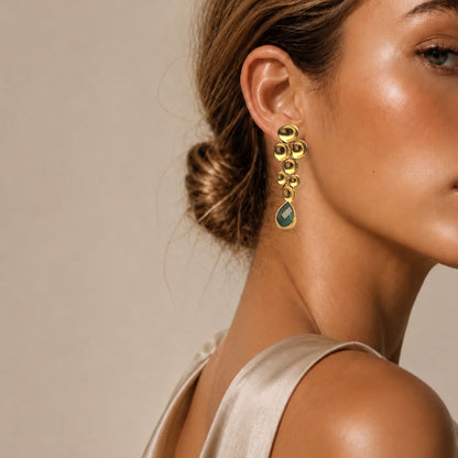 Woman wearing fancy sheer top and handmade gold earrings with green emerald gemstones against a neutral background