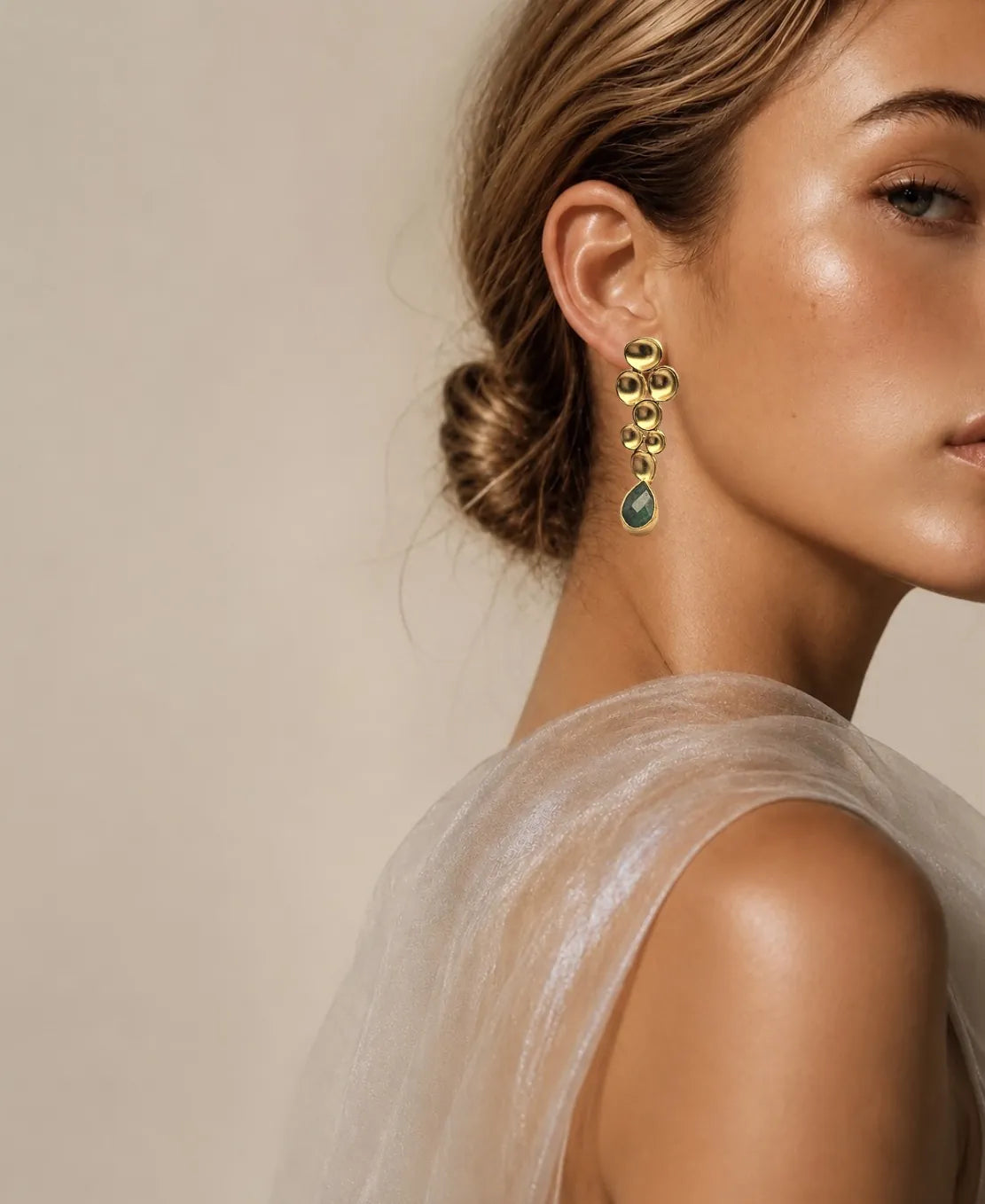 Woman wearing fancy sheer top and handmade gold earrings with green emerald gemstones against a neutral background