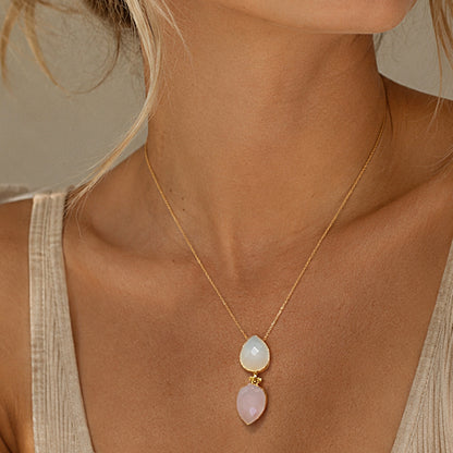 A close-up of a model wearing a delicate gold chain necklace with two teardrop-shaped pendants: one milky white snow Quartz and one light pink rose quartz paired with a beige ribbed tank top.