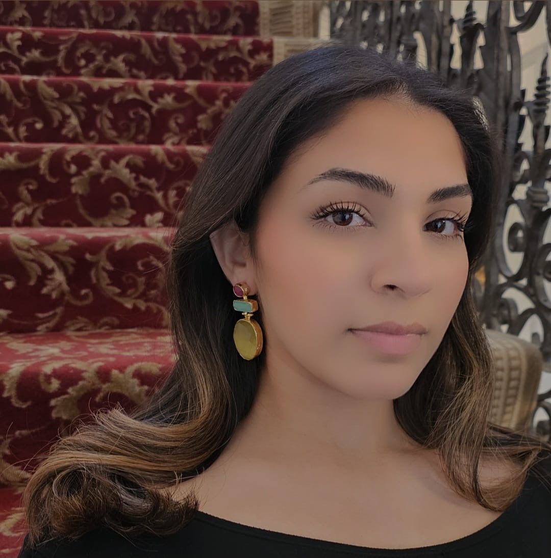 Model with hair curled sitting in mansion wearing A pair of gold drop earrings featuring a round ruby gemstone, a rectangular aqua chalcedony stone, and a large faceted oval yellow agate stone
