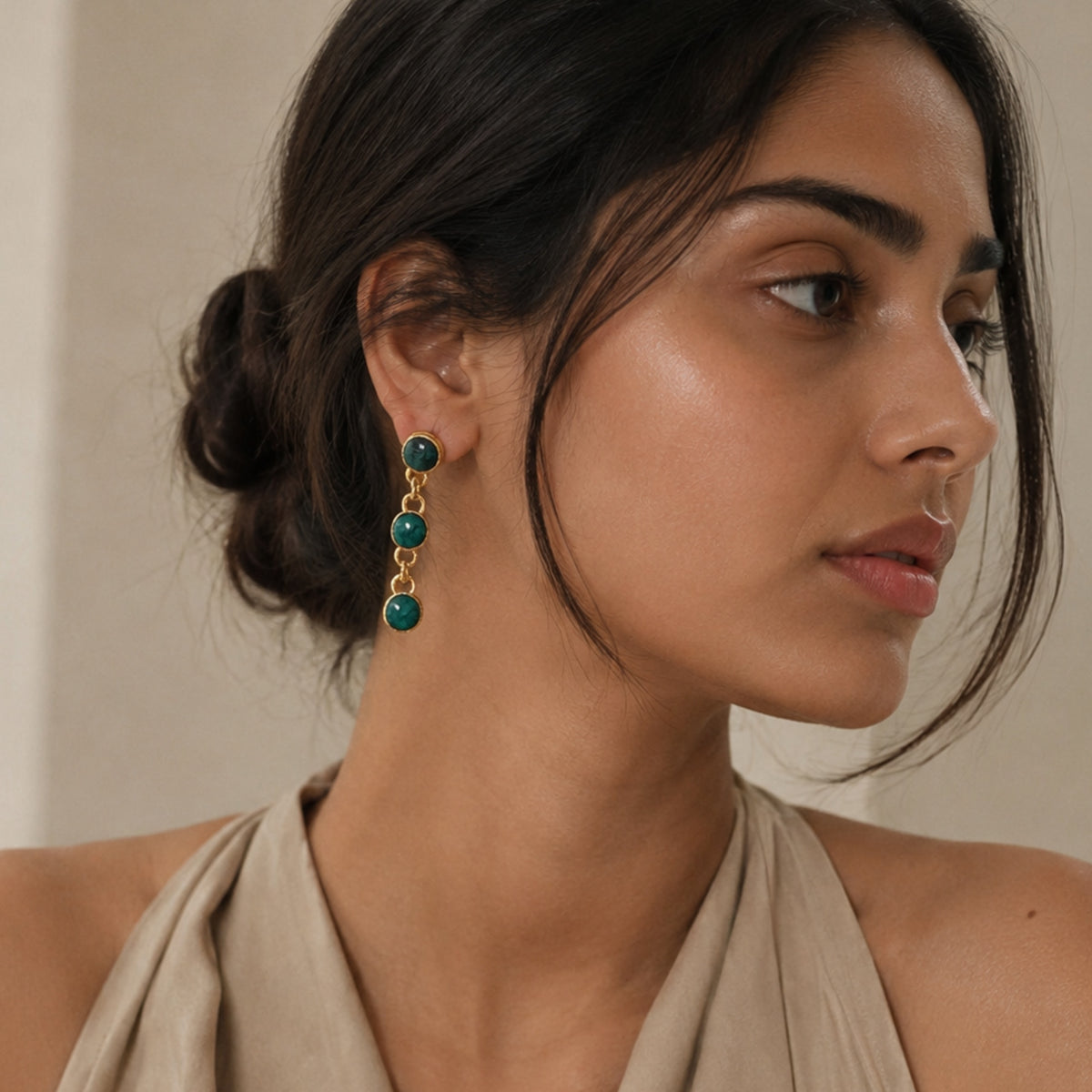 Model with low bun and beige halter top wearing Handmade gold-plated multi-stone emerald and chain dangle earrings on beige background 