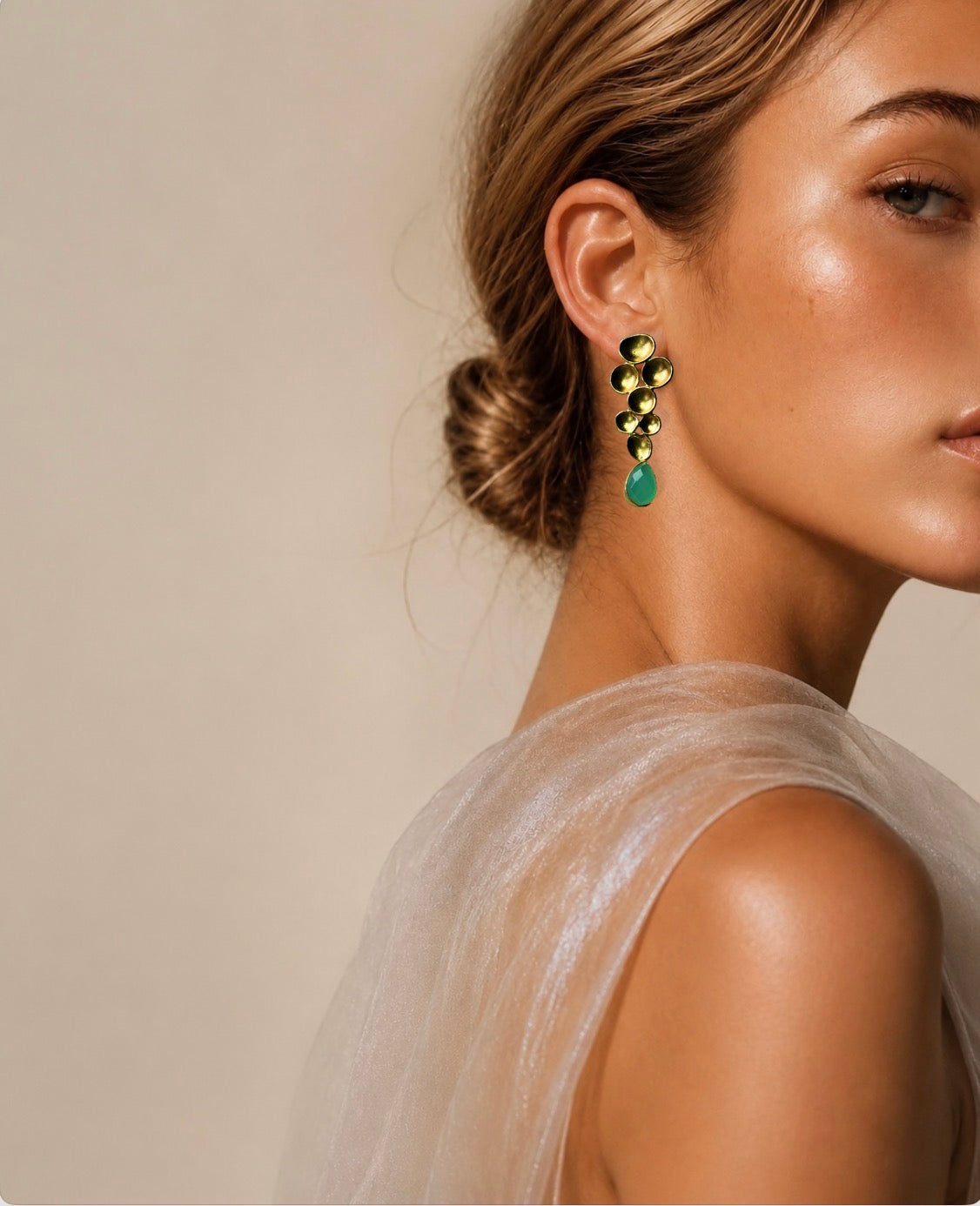 A close-up side profile of a woman wearing a contemporary gold-plated drop earring with clustered gold-toned oval stones and a teardrop-shaped aqua chalcedony gemstone, against a soft beige background.