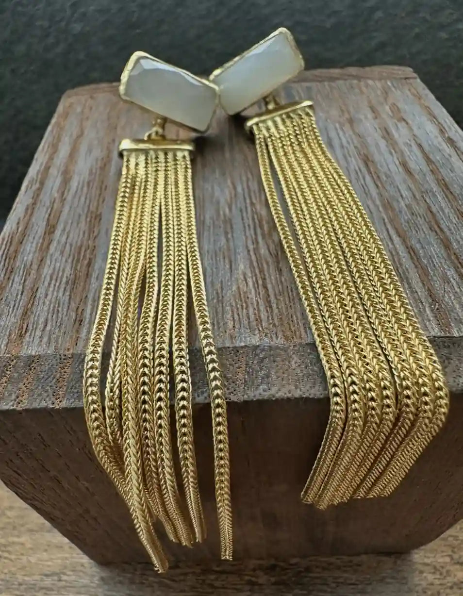 A pair of gold-toned statement earrings with rectangular cream-colored stone tops and long, multi-strand chain tassel, displayed on a wooden block.
