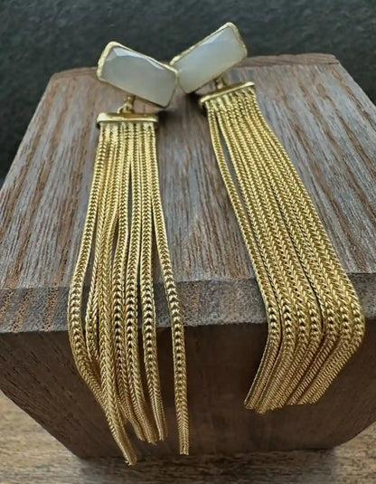 A pair of gold-toned statement earrings with rectangular cream-colored stone tops and long, multi-strand chain tassel, displayed on a wooden block.