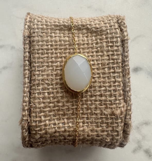 Minimal handmade gold-played chain bracelet with a snow Quartz pendant on a textured beige fabric against a marble background