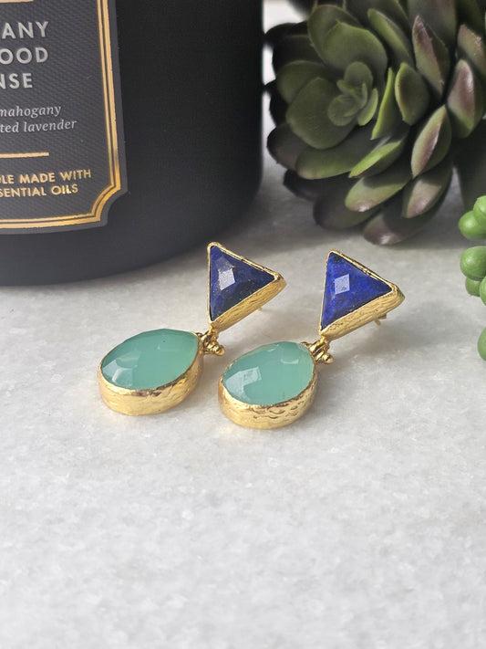 A pair of blue lapis and aqua chalcedony earrings with gold-plating over brass, displayed on a surface with a plant and a bottle in the background.
