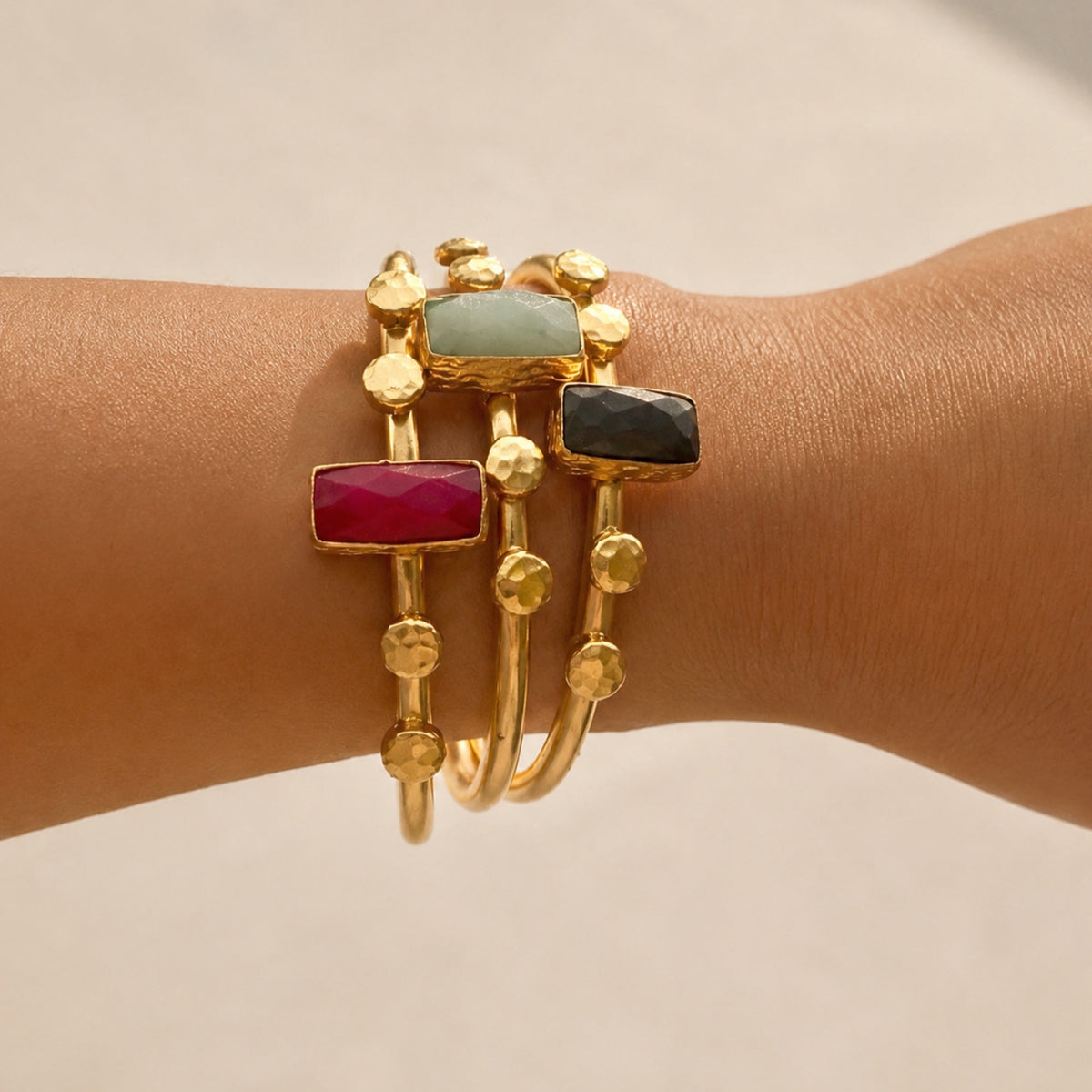 Three gold bangle bracelets with rectangular gemstone accents in ruby, lapis lazuli, and light gray, shown worn on a person's wrist against a warm beige background.