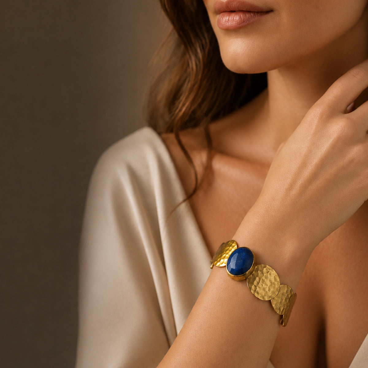 Woman in fancy dress wearing Gold-plated sustainable waterproof bangle with a blue lapis lazuli gemstone on a wrist against a brown background