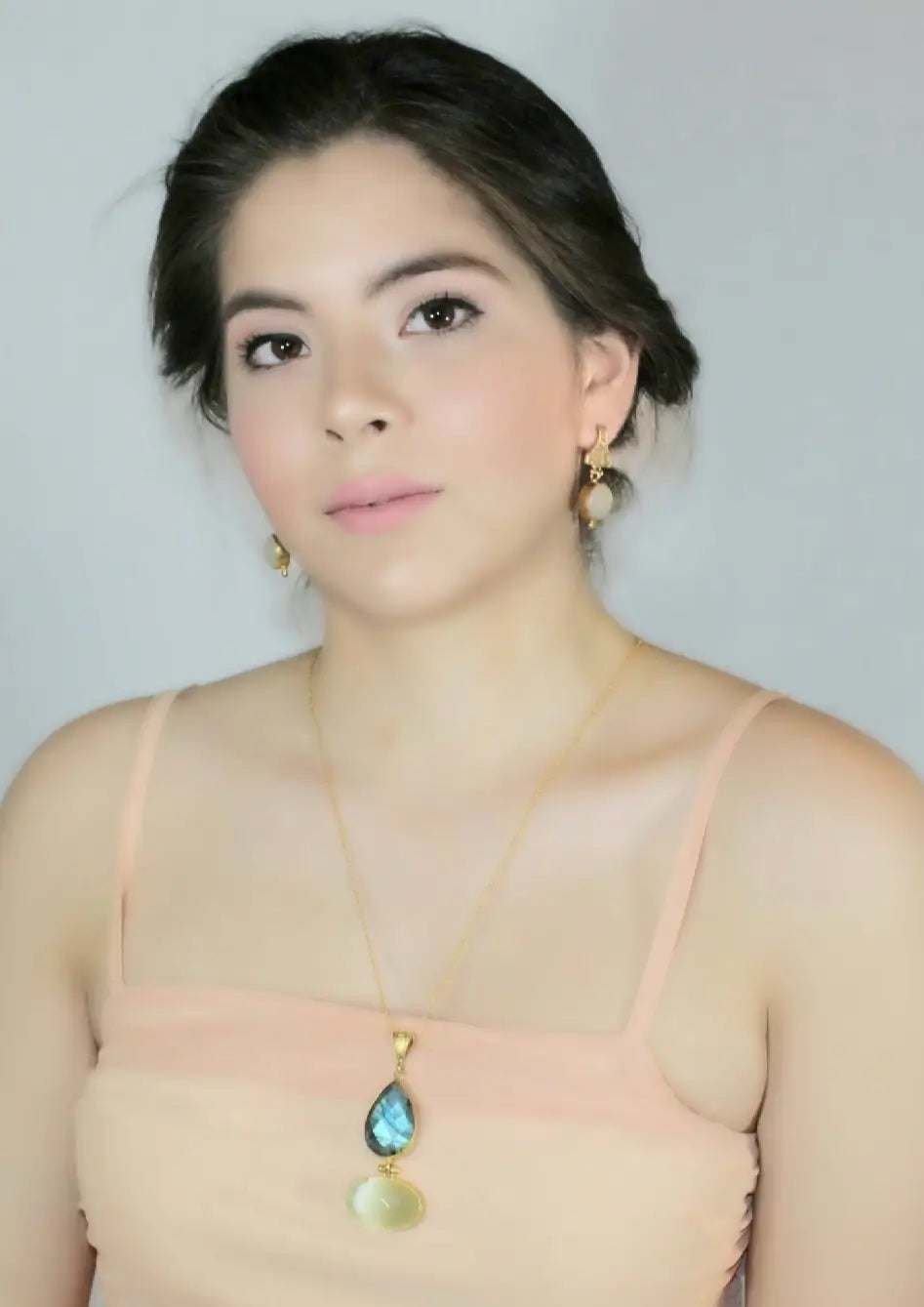 Woman wearing a beautiful long gold-plated necklace with a snow Quartz and labradorite pendant against a plain background