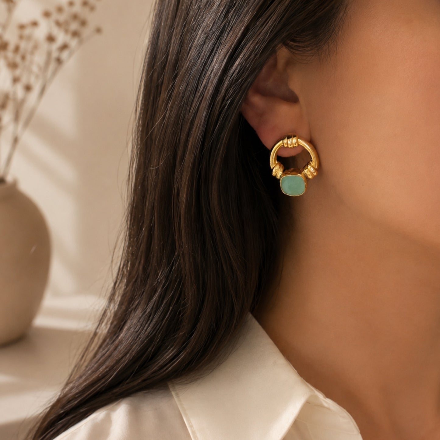 A close-up of a model's ear wearing a gold hoop earring with an aqua chalcedony center, set against a softly blurred background with a vase of dried flowers.
