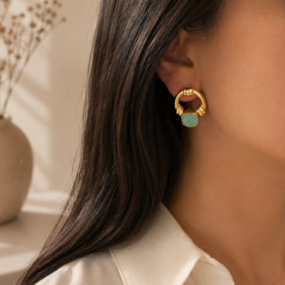 A close-up of a model's ear wearing a gold hoop earring with an aqua chalcedony center, set against a softly blurred background with a vase of dried flowers.