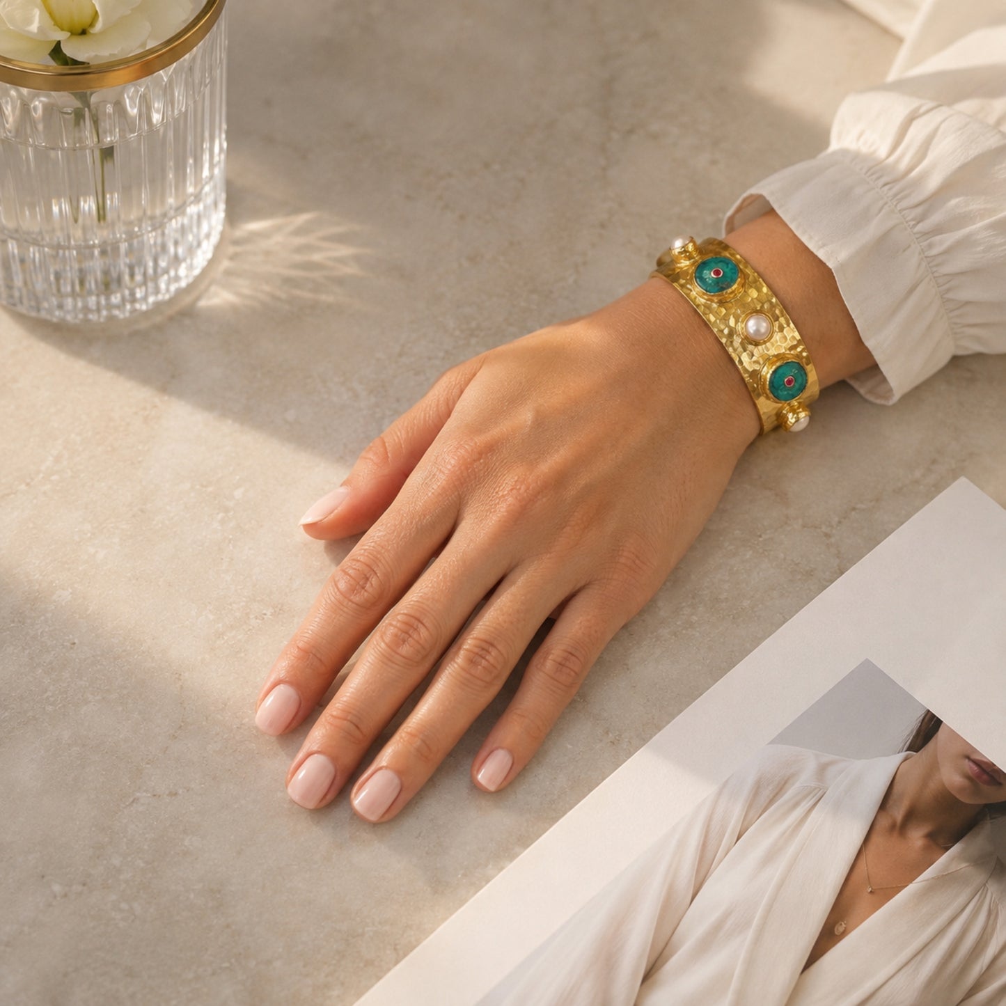 A woman’s hand showing her wrist wearing A handmade hammered gold cuff bracelet featuring turquoise inlay circles with small red zircon centers and white pearls shown on a neutral background