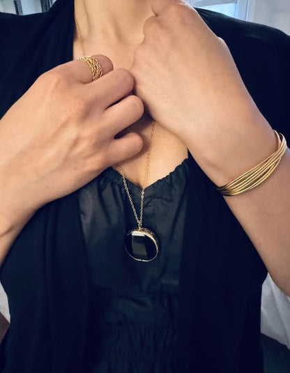 Woman’s torso wearing a black cardigan and jumpsuit with long, artisanal sustainable gold-plated round onyx pendant necklace hands grabbing chain.